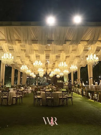 An expansive view of an elegant outdoor dining setup. I used a ceiling of white draped fabric and a grid of chandeliers to create a sense of a grand ballroom under the stars, with classic seating for a timeless feel.