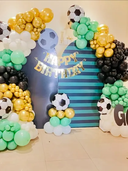 Score a goal with our football-themed birthday party decoration. This setup includes a goal-post-style balloon arch in green, black, gold, and white, with football foil balloons and a custom backdrop.