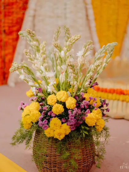 A rustic yet elegant floral arrangement in a woven basket, used as a table centrepiece. The mix of yellow marigolds and white tuberoses perfectly complements the traditional Haldi theme.