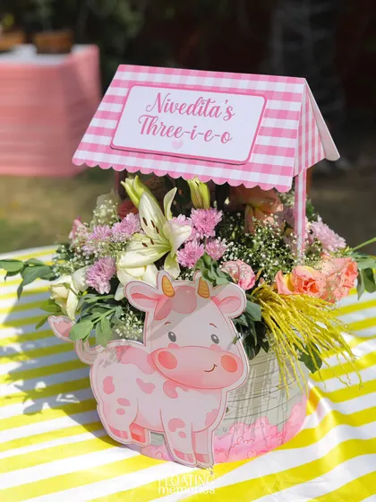 Our custom centerpieces featured fresh flowers in a basket, adorned with a pink gingham wishing well and a cute cow cutout. This detail carried the 'Three-i-e-o' farm theme to every table.