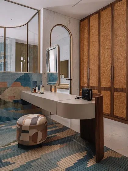 A detail of the vanity area in the master bathroom, with its curved wooden desk, patchwork pouf, and a view into the stunning cylindrical shower.