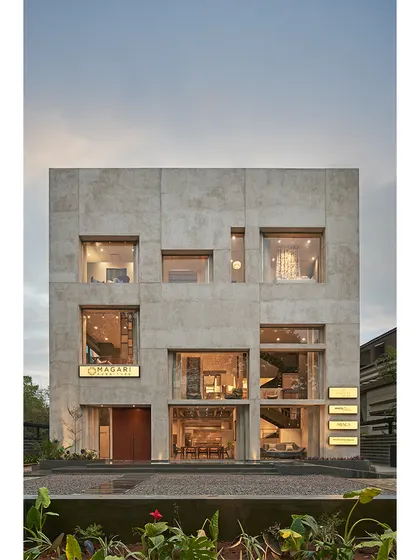 The facade of the Magari furniture store in Hyderabad features a monolithic form crafted from ferrocement with a sand plaster finish. Punched window openings create a dynamic composition and offer curated glimpses inside.