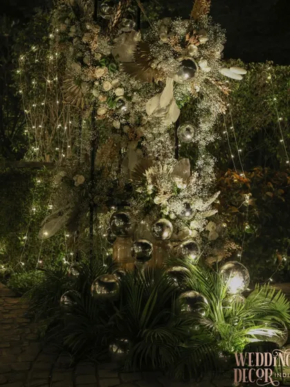 A stunning floral installation for an evening event, combining white flowers, dried palms, and silver baubles. The entire arrangement is interwoven with fairy lights, making it sparkle in the dark.
