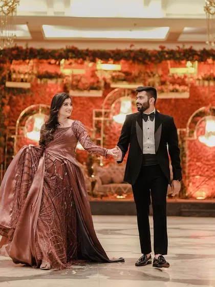 The couple walks hand-in-hand through their beautifully decorated reception hall. The bride's flowing gown and the red floral backdrop create a stunning visual.