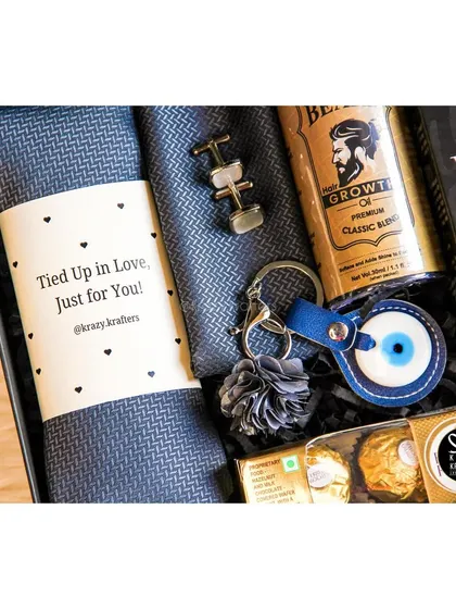 A close-up of the 'Styled with Love' hamper, showing the texture of the tie and the details on the evil eye keychain and cufflinks.