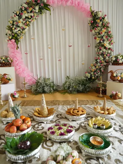 A full view of a half-saree ceremony setup, featuring a floral arch and traditional offerings beautifully arranged on silver platters. The pink fabric accents add a soft, celebratory touch.