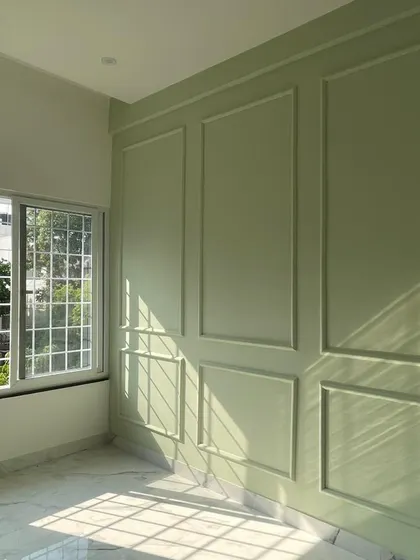 An on-site photo of a bedroom wall with freshly installed trim molding, painted in a calming pastel green. The natural light from the window highlights the clean lines and classic elegance of the design.