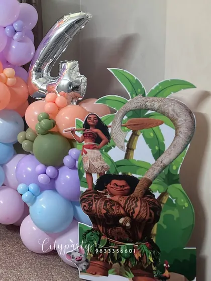 A 'Moana' themed birthday decoration for a 4th birthday. This setup includes character cutouts of Moana and Maui, a large '4' foil balloon, and a tropical-colored balloon garland.