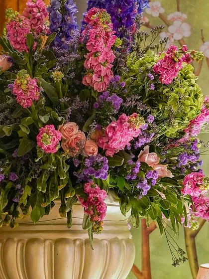 A large, lush floral arrangement in a classic white urn, featuring a beautiful mix of pink and purple blooms.