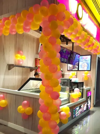 A custom balloon arch designed to fit the structure of a food kiosk for its opening day. The bright pink and yellow colors are cheerful and attract customers.