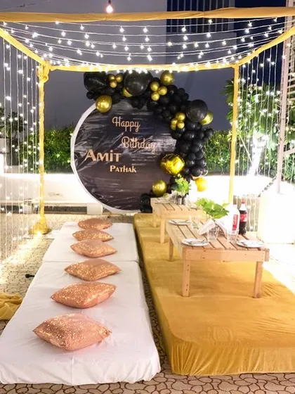 A cozy and stylish outdoor cabana setup for a birthday. The decor includes fairy lights, low seating with cushions, and a personalized backdrop with a black and gold balloon arch.