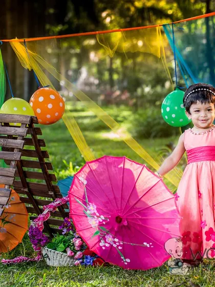 A little girl in a pink dress stands in a colorful outdoor setup with umbrellas and lanterns. The vibrant props and natural park setting make for a unique and joyful photo.