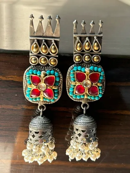 These fusion jhumkas are fit for royalty. They feature a crown-like top with intricate turquoise and red stone work, finished with a classic silver jhumka drop adorned with pearls.