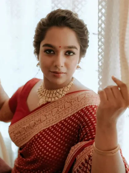 A candid portrait of a bride in a red saree, looking out of a window with a gentle smile.