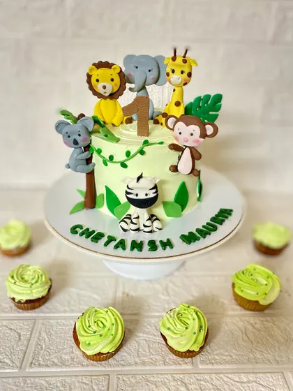 We designed this entire dessert setup for a first birthday party. It features a vanilla cake with hand-sculpted fondant jungle animals like a lion, elephant, and monkey, plus matching green-frosted cupcakes.