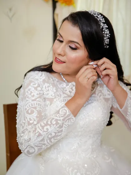 A candid moment of the bride putting on her earrings. Her hair is styled in long, flowing curls, and her makeup is a beautiful soft glam with a hint of shimmer.