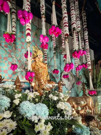 A close-up of the divine decor for a Krishna-themed event. A statue of Lord Krishna is surrounded by a sea of blue and white flowers, with garlands of pink and white flowers hanging above.