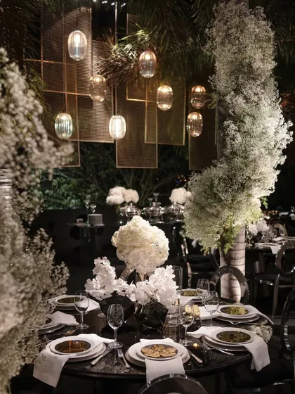 An intimate table setting at a glamorous evening event. The decor features dramatic baby's breath arrangements, hanging glass pendants, and a dark, moody color palette for a sophisticated look.