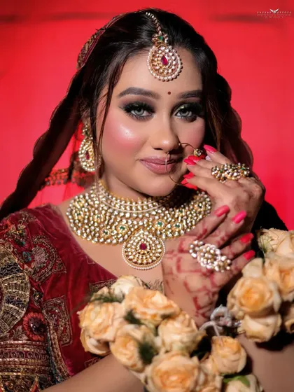A striking close-up of a bride in a deep red velvet lehenga. Her hands, adorned with floral garlands, frame her face, drawing attention to her bold eye makeup and heavy kundan necklace.