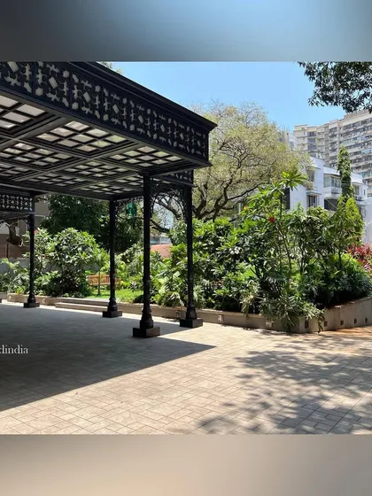 The ornate, dark metal structure of the covered entranceway contrasts beautifully with the bright, sunny garden beyond. The dappled shadows from the roof add another layer of texture to the paved ground below.