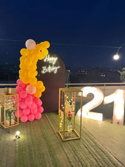 This is a duplicate of ID 21, 60, 67, 108, 121, 168, 206, 232, 239. A vibrant 21st birthday decoration on a rooftop. This setup features a balloon arrangement in hot pink and yellow, a 'Happy Birthday' neon sign, and large marquee numbers against a city skyline.