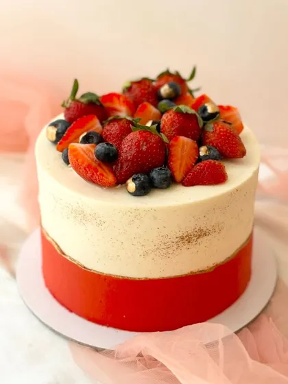 A striking two-tone cake with a red bottom half and a semi-naked top half. The cake is finished with a generous topping of fresh strawberries and blueberries.