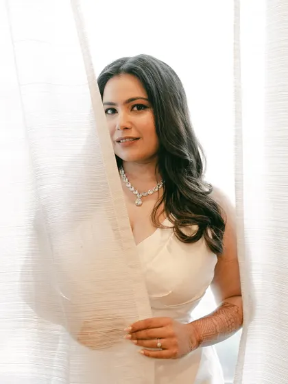 A playful and artistic shot. The bride peeks through the curtains, her makeup looking soft and radiant in the natural light.