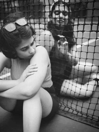 A black and white lifestyle shot of two women sitting on a tennis court, seen through the net. The image captures a candid, thoughtful moment of friendship and connection.