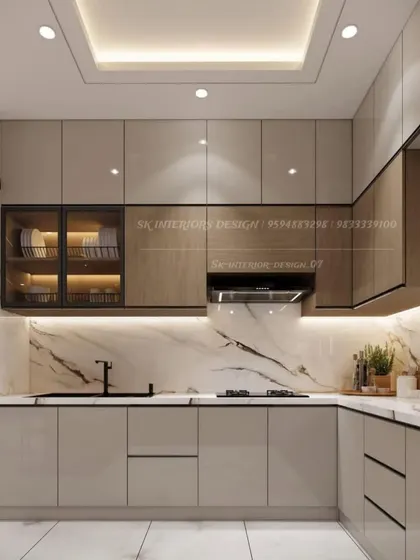 A luxurious L-shaped kitchen combining beige and wood-finish cabinets with a stunning marble backsplash. The design includes a glass-fronted cabinet and a false ceiling with integrated lighting.