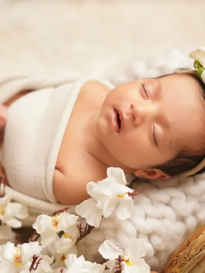 A newborn girl sleeping peacefully, surrounded by white flowers. The soft, angelic styling with a floral headband creates a truly timeless and beautiful infant portrait.