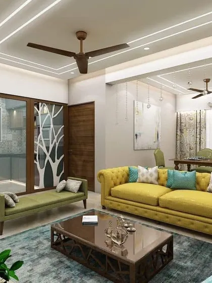 A vibrant living room with a yellow tufted sofa and blue accents. The tree-motif partition separating the kitchen adds a creative and artistic element.