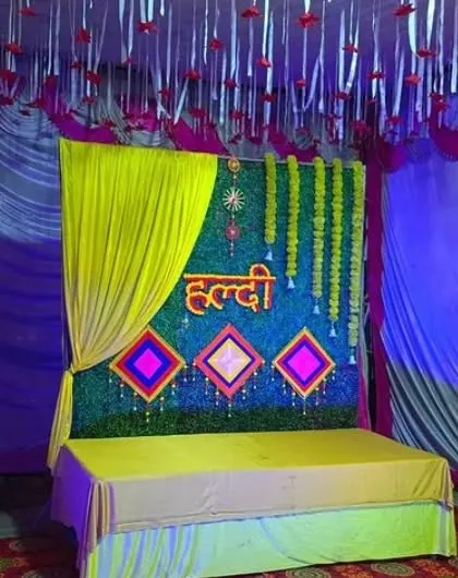 A Haldi stage with a green grass backdrop, yellow draping, and a decorated ceiling with hanging ribbons.
