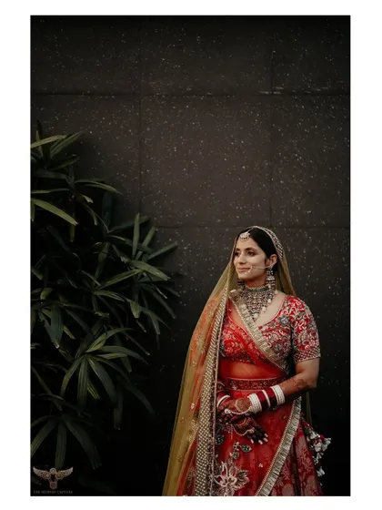 A classic bridal portrait against a dark, minimalist background. The lighting is focused entirely on the bride, making her red and gold lehenga appear even more vibrant and rich.