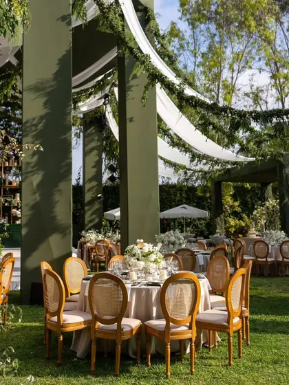 A round dining table setup in the garden. The combination of the neutral tablecloth, wooden cane-back chairs, and a simple white floral centerpiece creates a timeless and elegant look.