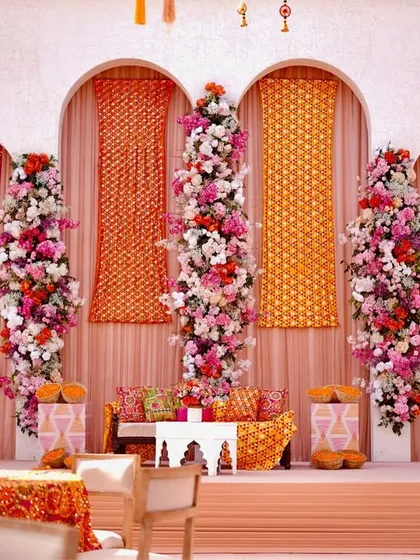 The stage for the Phulkari Mehendi, featuring a backdrop of white arches, floral pillars, and panels of traditional 'phulkari' fabric. The seating is adorned with colorful embroidered cushions.