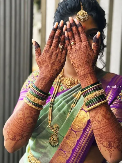 A shy bride peeking through her hands, which are adorned with a beautifully dark mehndi stain. The rich color enhances the intricate jaali (net) pattern.