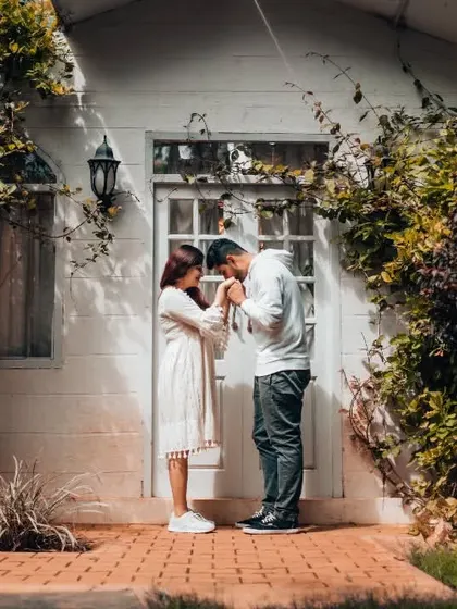 A full-length shot of a couple sharing a tender moment in front of a white cottage, framed by lush greenery. A perfect pre-wedding setting.