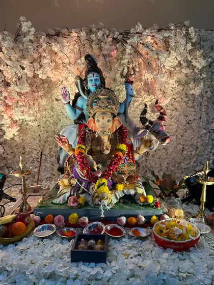 An elegant and serene home Ganpati setup featuring a beautiful idol of Ganesha with Shiva and Parvati. The backdrop is a dense wall of delicate white blossoms, creating a heavenly and peaceful ambiance for prayer and offerings.