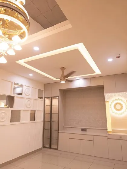 This living area is a masterpiece of modern luxury. A stunning chandelier hangs from a mirrored ceiling panel, complemented by a custom TV unit and an elegant pooja space, all unified by a clean white and gold theme.