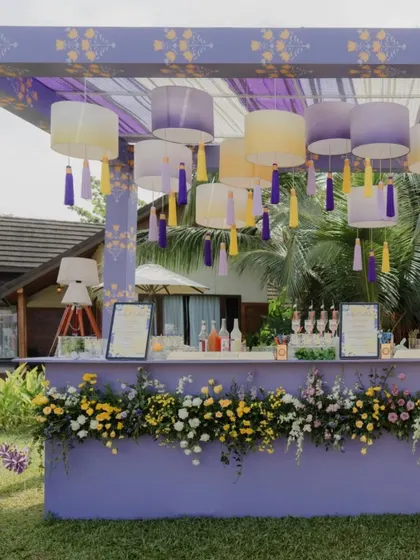 A chic outdoor cocktail bar designed for a pastel-themed Haldi. The lavender-colored bar is adorned with fresh flowers and features an overhead canopy with hanging lampshades and colorful tassels.