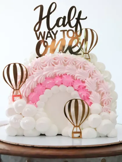 A close-up of the 'Half Way to One' cake, showing the texture of the piped rainbow and the cute gold hot air balloons.