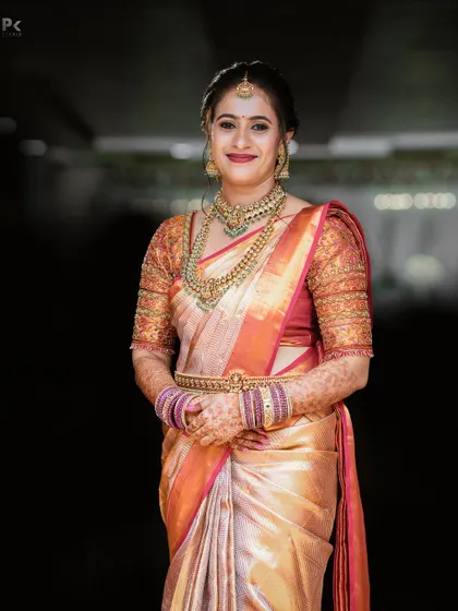 A full-length portrait of a bride in a stunning Kanjeevaram saree. Her makeup is bold and beautiful, with a bright lip color that stands out against the rich fabric and heavy gold jewellery.
