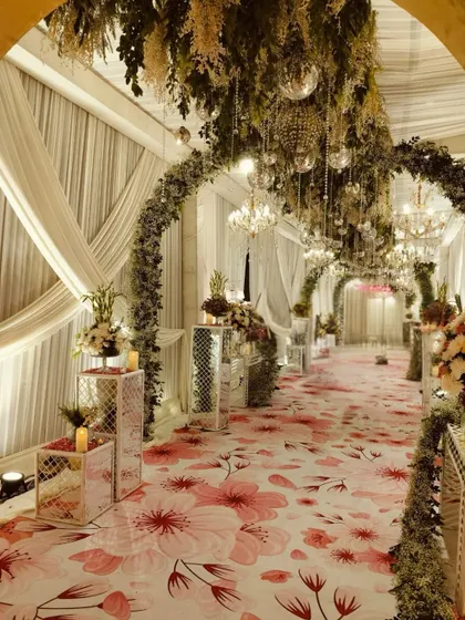 A fairytale passage. The aisle is lined with greenery-filled arches and lit by sparkling chandeliers, with a beautiful floral-patterned carpet underfoot.