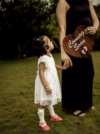A touching moment between mother and daughter. The little girl holds a "Coming Soon" sign, looking up at her mom and the new baby bump with curiosity and love.