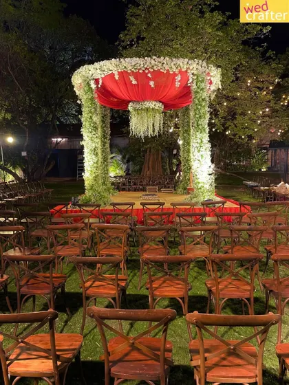 A bold and contemporary mandap for an outdoor night ceremony. The structure features pillars of lush greenery, a vibrant red canopy, and a dramatic chandelier of white flowers.