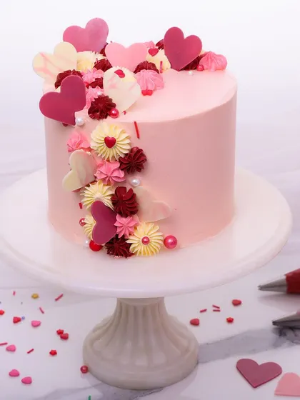 We craft a cascade of love with this romantic anniversary cake. A beautiful arrangement of red, pink, and white chocolate hearts and buttercream flowers flows down the side of a soft pink cake, creating a design that is bursting with affection.