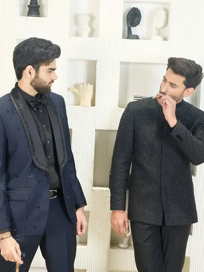 An interactive moment between two gentlemen, one wearing a navy tuxedo with an embroidered shawl collar and the other in a black Jodhpuri. Both outfits are designed for sophisticated evening events.