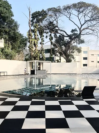 A swimming pool with a bold, black-and-white checkerboard pattern, creating a surreal, graphic landscape that reflects the sky and surrounding trees.