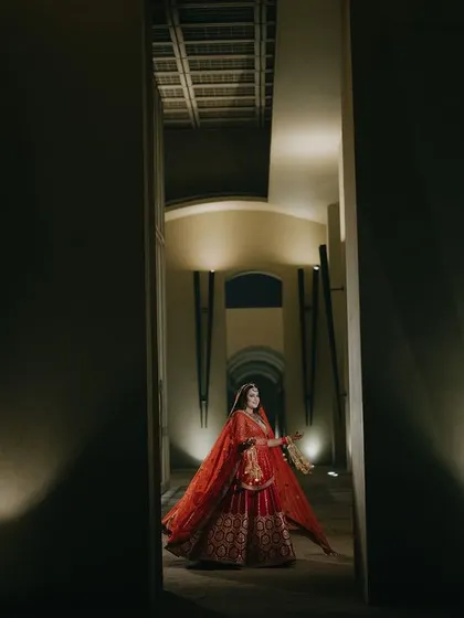 A grand portrait of Tamanna in her red bridal lehenga, framed by a dark corridor. The lighting highlights her, creating a dramatic and powerful image that emphasizes her stunning attire.