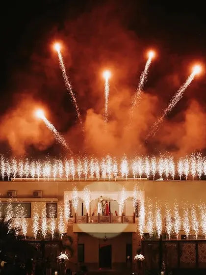 A grand, wide shot of a palace wedding at night, with fireworks illuminating the entire building as the couple stands on the balcony.